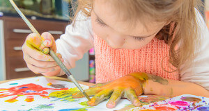 Young female child painting her hand on top of a desk of paintings.